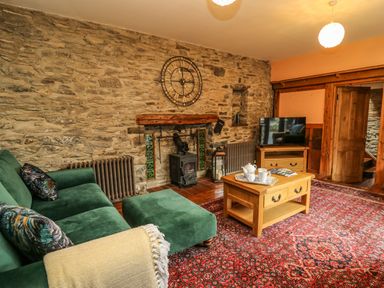 A living room with a green sofa and ottoman a wooden coffee table on a patterned red rug a stone wall with a clock and fireplace a television on a wooden cabinet at Pant Y Rhedyn in Abbeycwmhir near Rhayader