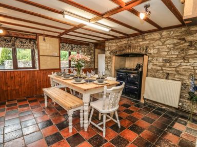 A dining room with a wooden table set for six with bowls glasses and a flower vase with benches and chairs by a stone wall and a traditional stove at Pant Y Rhedyn in Abbeycwmhir near Rhayader