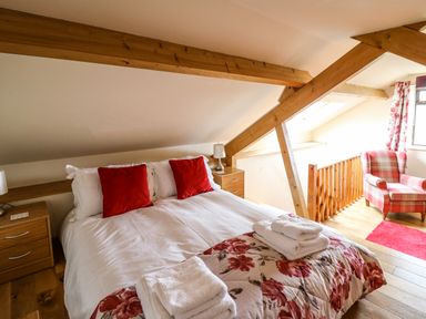 A bedroom with a double bed with red pillows and floral bedding next to wooden beams and a plaid armchair at Pant Y Rhedyn in Abbeycwmhir near Rhayader