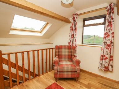 A stair landing with a plaid armchair wooden railing a skylight and a window with floral curtains at Pant Y Rhedyn in Abbeycwmhir near Rhayader