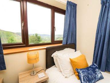 A bedroom with a bed and pillows next to a window with blue curtains and a lamp on a wooden nightstand at Pant Y Rhedyn in Abbeycwmhir near Rhayader