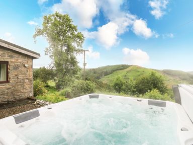 An outdoor hot tub with bubbling water next to a stone building and a view of green hills and trees at Pant Y Rhedyn in Abbeycwmhir near Rhayader
