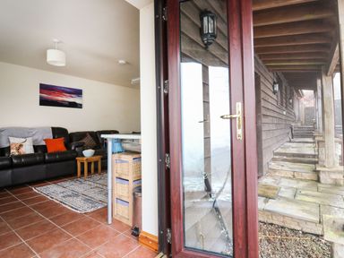 A living room with a black leather sofa and colorful cushions next to a glass door opening to a stone porch at Pant Y Rhedyn in Abbeycwmhir near Rhayader