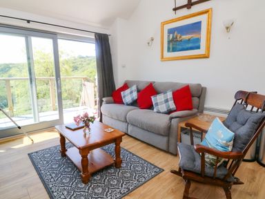 A living room with a gray sofa with red and checkered cushions a wooden rocking chair a wooden coffee table on a patterned rug and sliding glass doors at Higher Bumsley Barn in Parracombe