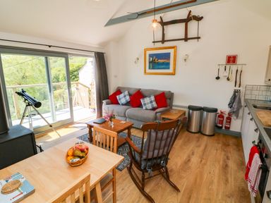 A living room with a grey sofa and red cushions a rocking chair a wooden dining table with fruit on it and a telescope near a sliding glass door at Higher Bumsley Barn in Parracombe