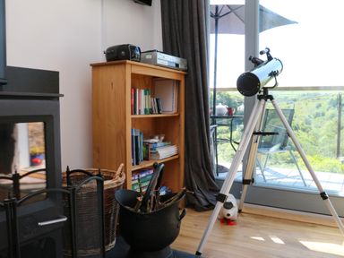 A wooden bookshelf with books and devices next to a fireplace and a telescope by a glass door at Higher Bumsley Barn in Parracombe