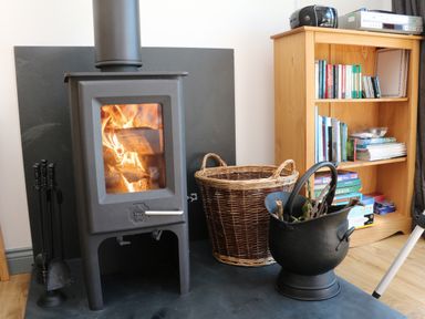 A burning wood stove with fireplace tools a wicker basket and a black bucket with sticks next to a wooden bookshelf at Higher Bumsley Barn Parracombe