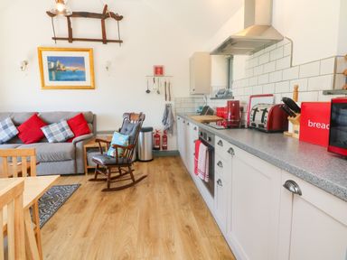 A kitchen and living area with a gray sofa and red and blue cushions a wooden rocking chair a kitchen counter with appliances and a wooden floor at Higher Bumsley Barn Parracombe