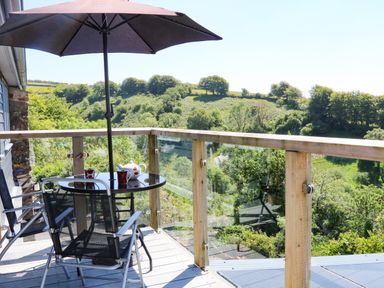 An outdoor balcony with a glass railing a table with an umbrella two chairs and a teapot and cups overlooking a green landscape at Higher Bumsley Barn Parracombe