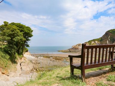 A wooden bench overlooking a rocky beach and sea with trees and cliffs in the background at Higher Bumsley Barn Parracombe