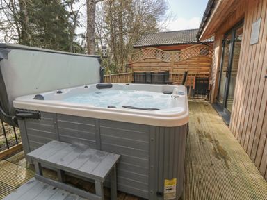 An outdoor hot tub on a wooden deck next to a wooden building with patio furniture and trees in the background at Treetops in Otterburn