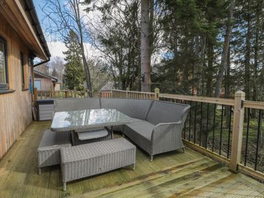 An outdoor wooden deck with a wicker corner sofa and glass top table surrounded by trees at Treetops in Otterburn