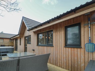 An outdoor patio area with a glass table and wicker chairs next to a wooden building with windows and a hot tub at Treetops in Otterburn