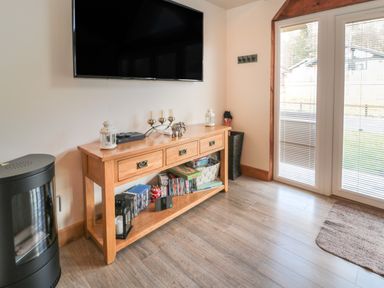 A wooden console table with decorative items and DVDs under a wall mounted TV next to glass doors with blinds at Treetops in Otterburn