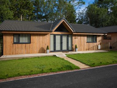 A wooden cabin with a black tiled roof and glass doors surrounded by grass and trees at Treetops in Otterburn
