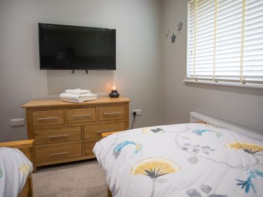 A bedroom with two single beds floral bedding a wooden chest of drawers with folded towels and lamp a TV mounted on the wall white window blinds at Treetops in Otterburn