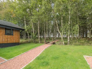 A wooden building beside a gravel path with a small bridge leading into a wooded area at Treetops in Otterburn