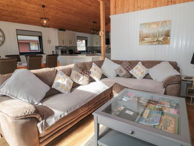 A living room with a large sectional sofa and glass coffee table showing board games with a view into a kitchen with dining chairs at Pine Tree in Otterburn