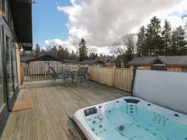 An outdoor wooden deck with a hot tub and a table with chairs surrounded by wooden houses and trees at Pine Tree in Otterburn