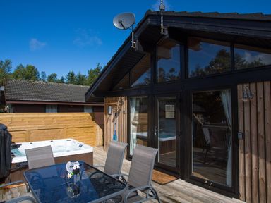 An outdoor patio with a glass table and four chairs a hot tub wooden fence and a wooden house with large windows at Pine Tree in Otterburn