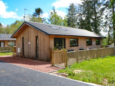 A wooden single-story house with a fenced patio and trees in the background at Broken-Sky Lodge in Otterburn