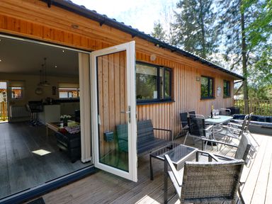 An outdoor wooden deck with seating and a table next to a wooden building with open glass doors at Broken-Sky Lodge in Otterburn