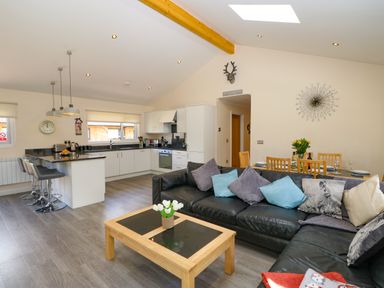 An open plan living area with a black leather sofa and a wooden coffee table with flowers a kitchen with white cabinets and a breakfast bar with stools and a dining table with wooden chairs at Broken-Sky Lodge in Otterburn