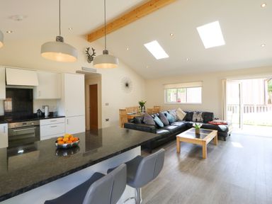 An open plan kitchen and living area with a black countertop island and stools a dining table and a black corner sofa at Broken-Sky Lodge in Otterburn