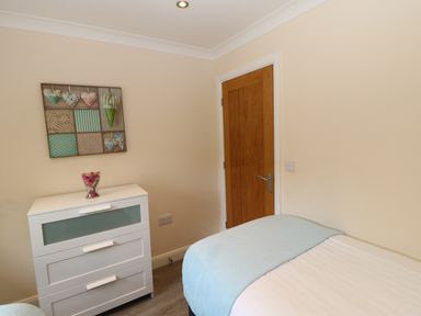 A bedroom with a white chest of drawers a bed with a light blue blanket a wooden door and wall art at Broken-Sky Lodge in Otterburn