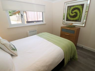 A bedroom with a bed covered by white sheets and a green blanket next to a wooden dresser with a framed picture above at Broken-Sky Lodge in Otterburn