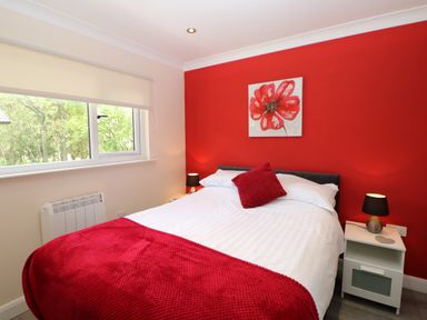 A bedroom with a double bed red blanket and cushion red accent wall with flower painting and bedside tables with lamps at Broken-Sky Lodge in Otterburn