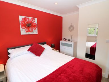 A bedroom with a double bed white and red bedding red wall painting a white chest of drawers and a wall mirror at Broken-Sky Lodge in Otterburn