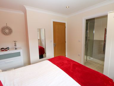 A bedroom with a bed covered in white and red bedding a wooden door a white dresser with decor and a door to a tiled bathroom with a glass shower at Broken-Sky Lodge in Otterburn