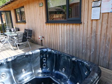 An outdoor area with a black hot tub wooden deck a glass table with four chairs and a wooden building wall with windows at Broken-Sky Lodge in Otterburn