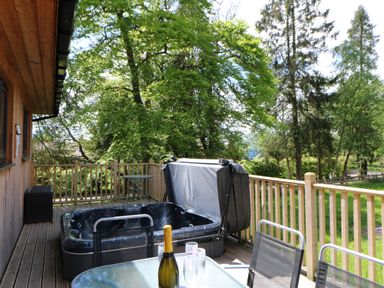 An outdoor wooden deck with a hot tub a glass table with a bottle and glasses and metal mesh chairs at Broken-Sky Lodge in Otterburn