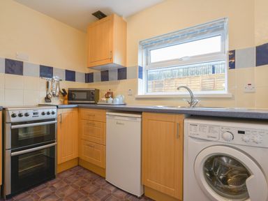 A kitchen with oven microwave small refrigerator washing machine and wooden cabinets at Broadclyst Cottage in Ottery St Mary