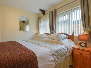 A bedroom with a double bed beside a wooden bedside table with a lamp and windows with curtains at Broadclyst Cottage in Ottery St Mary