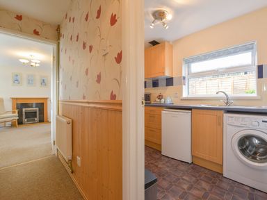A hallway with patterned wallpaper leading to a living room with a chair and fireplace and a kitchen with cabinets washing machine and a window at Broadclyst Cottage in Ottery St Mary