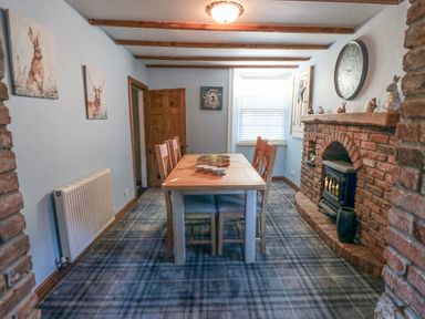 A dining room with a wooden table and six chairs a brick fireplace with a stove and a large wall clock at The Nook at 29 High Street Cloughton near Burniston