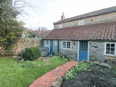 A stone cottage with blue doors and windows a brick pathway and garden in front at The Nook at 29 High Street in Cloughton near Burniston
