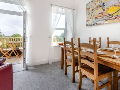 A dining area with a wooden table set for four and wooden chairs next to a window and open door leading to a balcony with a table and chairs at Barwood View in Torquay