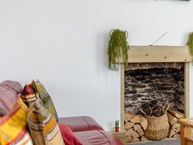 A fireplace with stacked firewood and a basket of pinecones next to a red sofa with colorful cushions at Barwood View in Torquay