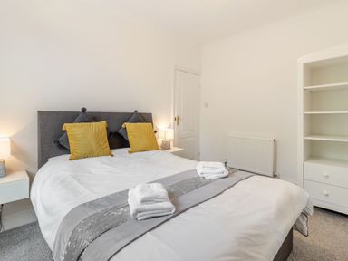 A bedroom with a double bed yellow and grey pillows bedside tables with lamps and a white shelving unit at Barwood View in Torquay