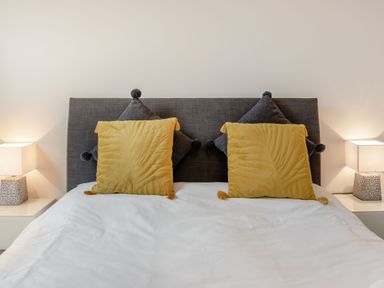 A bedroom with a bed with a grey headboard two sets of pillows and two bedside tables with lamps at Barwood View in Torquay
