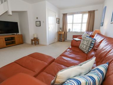 A living room with an orange leather sofa pillows a wooden TV stand with a television a small side table lamp and window with curtains at Fairways in Wyke Regis