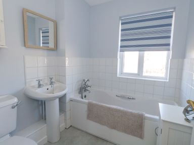 A bathroom with a bathtub a sink a toilet a window with striped blinds and a small cabinet at Fairways in Wyke Regis