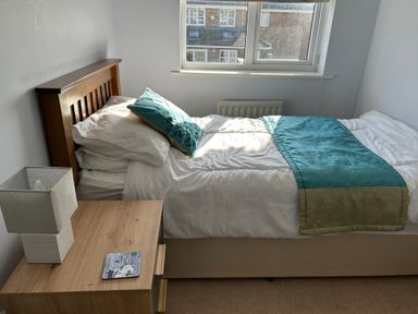 A bedroom with a single bed with blue and beige bedding and a wooden nightstand with a lamp and coaster near a window at Fairways in Wyke Regis