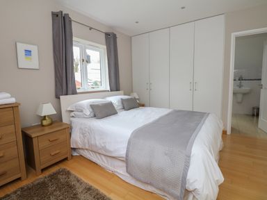 A bedroom with a double bed white bedding gray pillows and throw a wooden nightstand with a lamp a window with gray curtains white built-in wardrobes wooden flooring and a door leading to a bathroom at Cholwell Barn Apartment in Mary Tavy