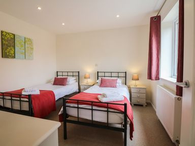 A bedroom with two single beds with red covers and pillows two bedside tables with lamps a window with red curtains and a radiator at Cholwell Barn Apartment in Mary Tavy