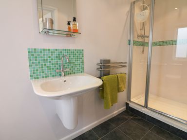 A bathroom with a white sink green tiled backsplash glass shelf soap bottles towel rack with green towels and a glass shower enclosure at Cholwell Barn Apartment Mary Tavy